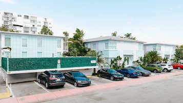 Un parking rempli de voitures, une clôture verte et des bâtiments aux façades bleu clair.