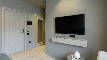 Een moderne hotelkamer met een flatscreen televisie aan de muur, een klein rond tafeltje met een boek en een vaas, en een grijze bank.