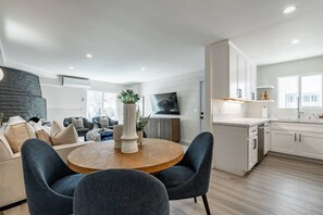 Une cuisine moderne avec des meubles blancs, un coin repas avec une table ronde et des chaises, et un salon avec un canapé et des coussins décoratifs.