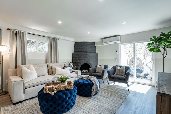 Un salon moderne avec un canapé, des poufs, une cheminée et une grande fenêtre.
