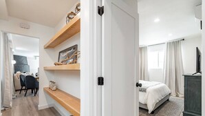 A hotel room with a bed, a desk, and a chair. There is a shelf with decorative items and a basket with towels. - The Rosemont - La Jolla Luxury (La Jolla)