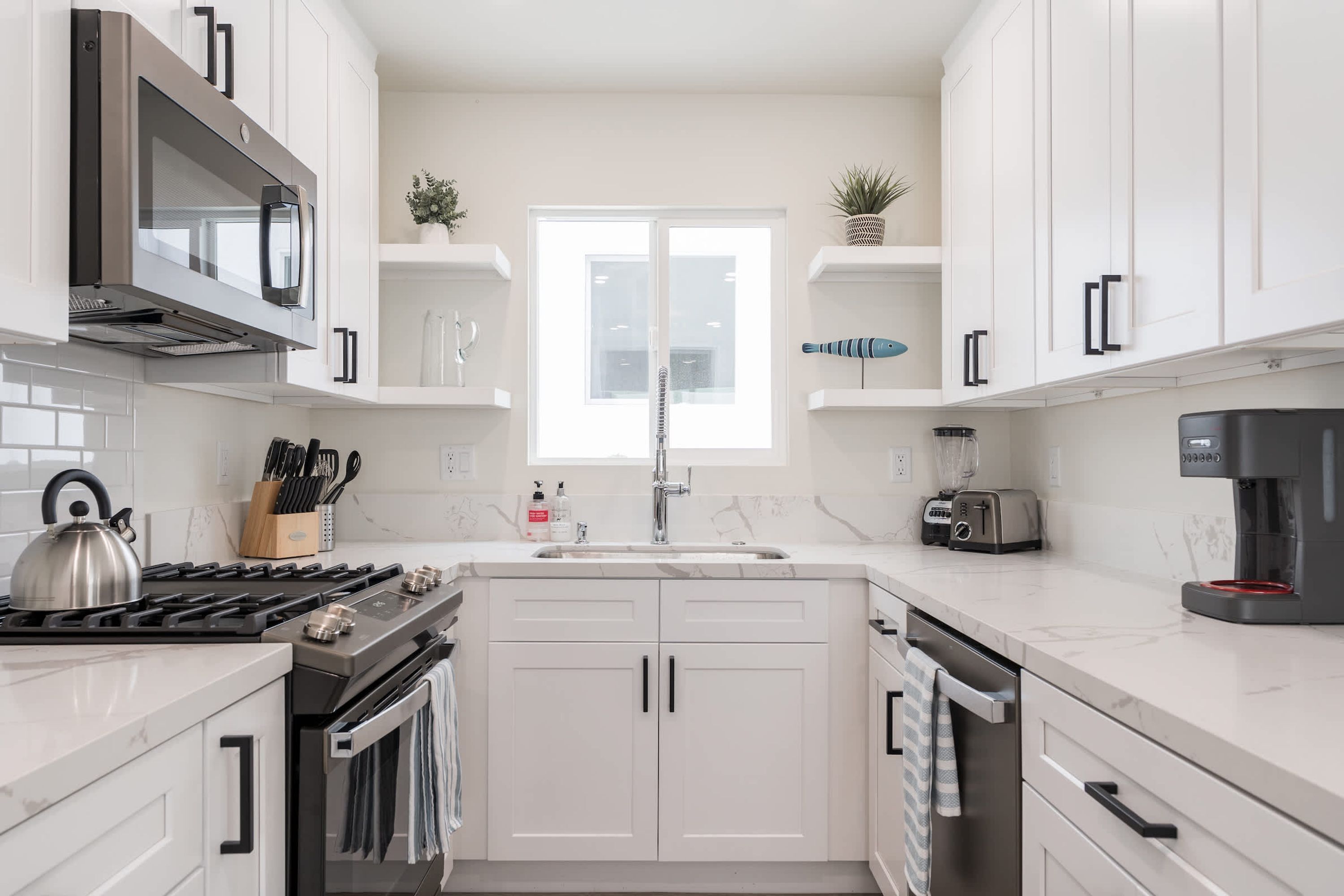 Una cocina moderna con armarios blancos, una estufa de acero inoxidable, horno empotrado, un lavamanos y una cafetera.