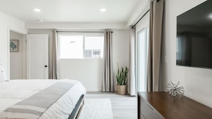 Un dormitorio moderno con una cama grande, una mesita de noche, un televisor montado en la pared y una ventana con cortinas.