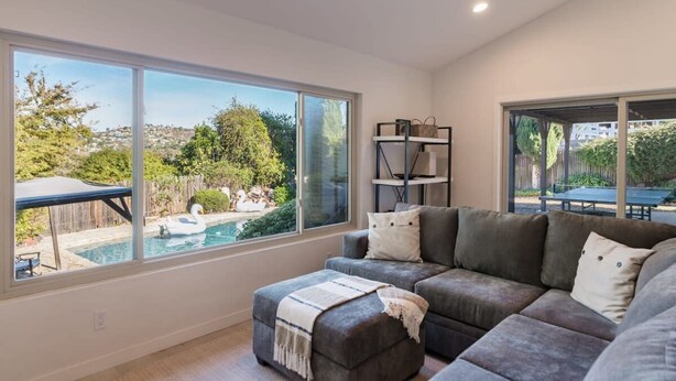 A modern living room with a large window, a grey sectional sofa, and a small ottoman. - Spacious and Bright 5-bedroom Oasis Pool and Yard (La Mesa)