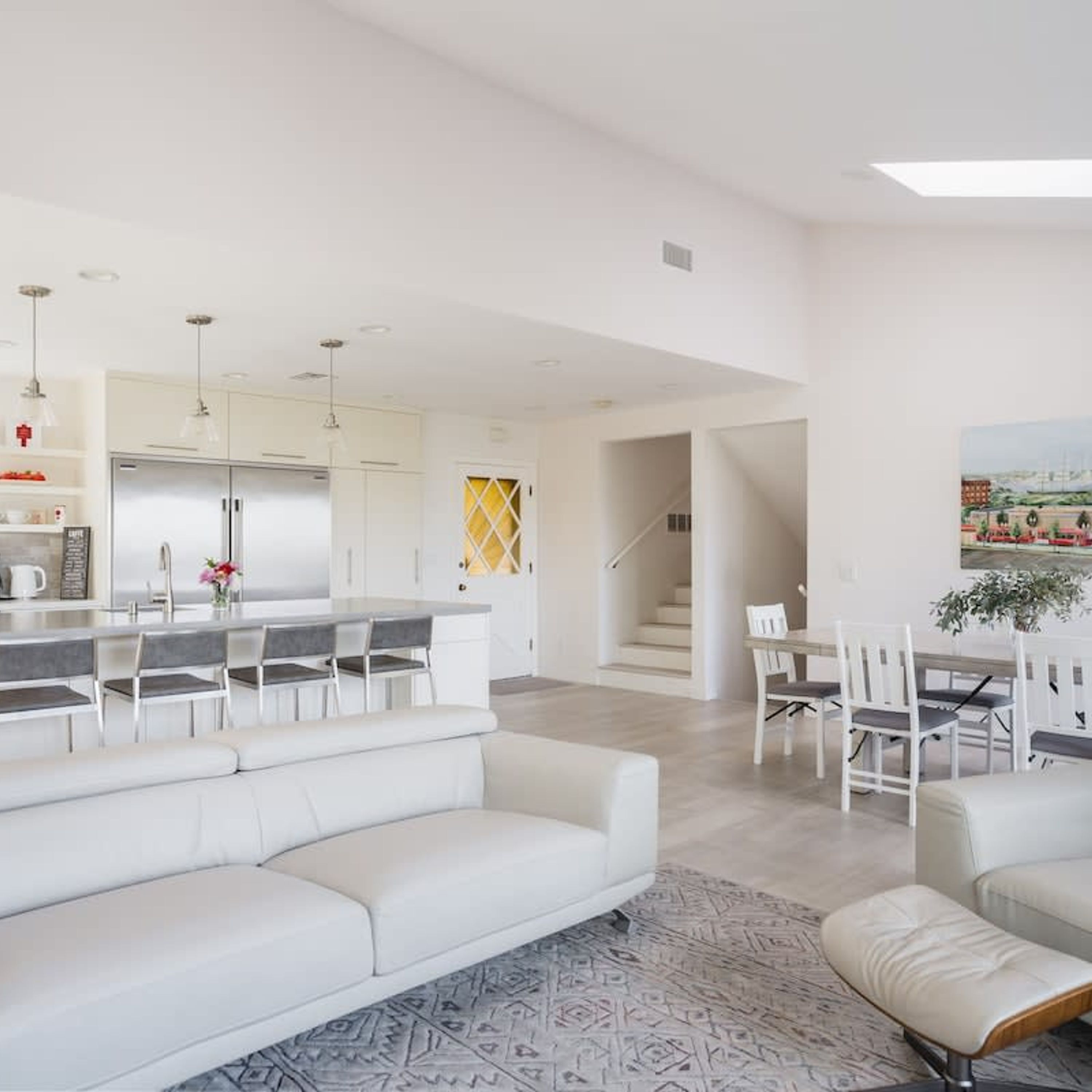 A modern living room with a white sofa, a dining area with white chairs, and a kitchen with white cabinets and a stainless steel refrigerator.