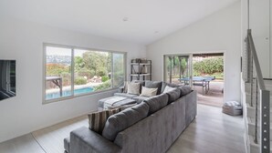 A spacious living room with a large grey sectional sofa, a flat-screen TV, and a view of a swimming pool and outdoor furniture. - Spacious and Bright 5-bedroom Oasis Pool and Yard (La Mesa)