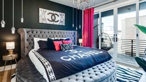A bedroom with a tufted headboard, a bed with a Chanel-themed bedspread, a chandelier, a nightstand with a lamp, and a view of the outside balcony. - Music Fame House Gulch 4 Patios View Karaoke (Nashville)