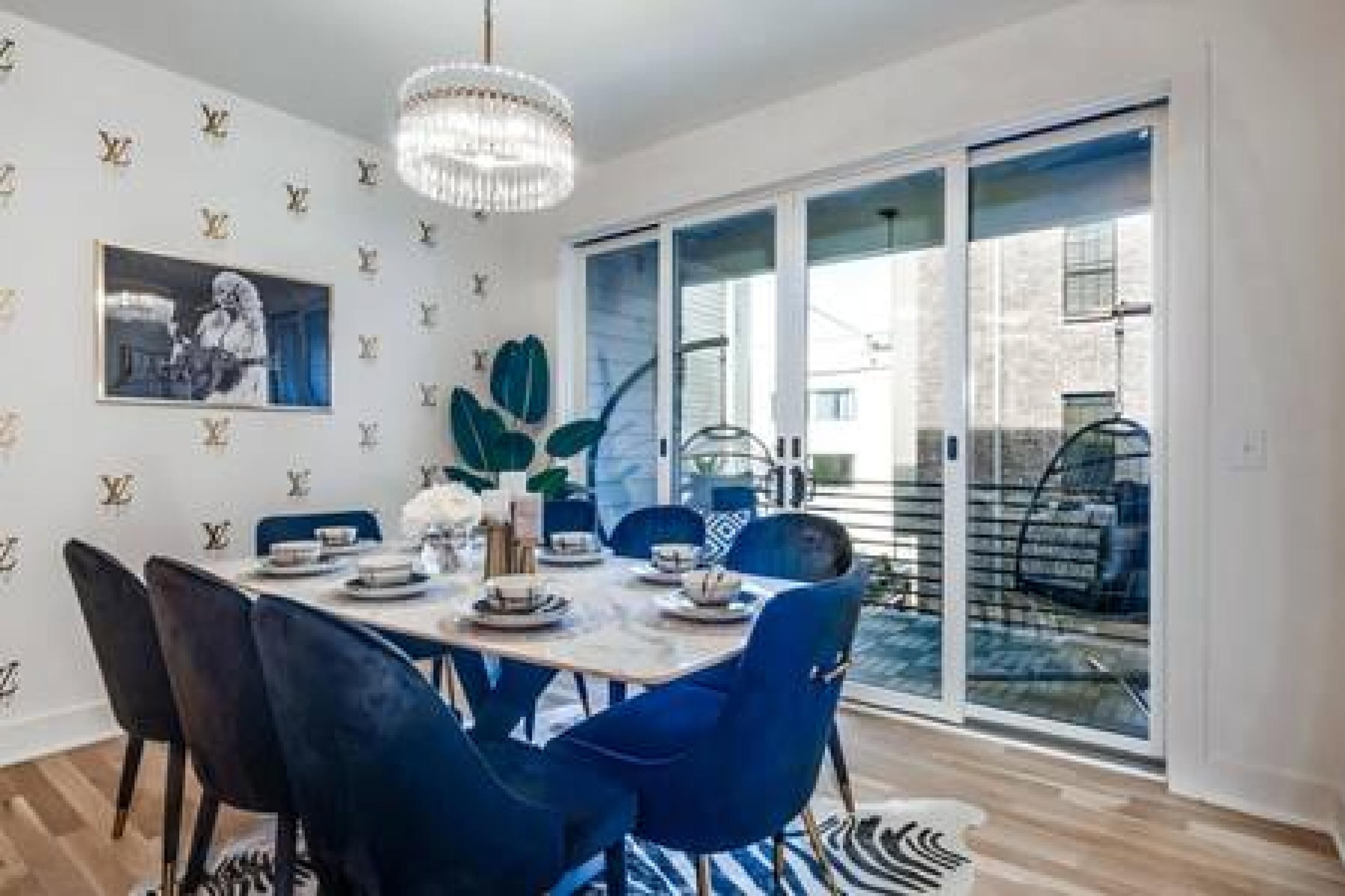Un coin repas avec une table dressée pour quatre personnes, des chaises bleues, un lustre et une grande fenêtre donnant sur l’extérieur.