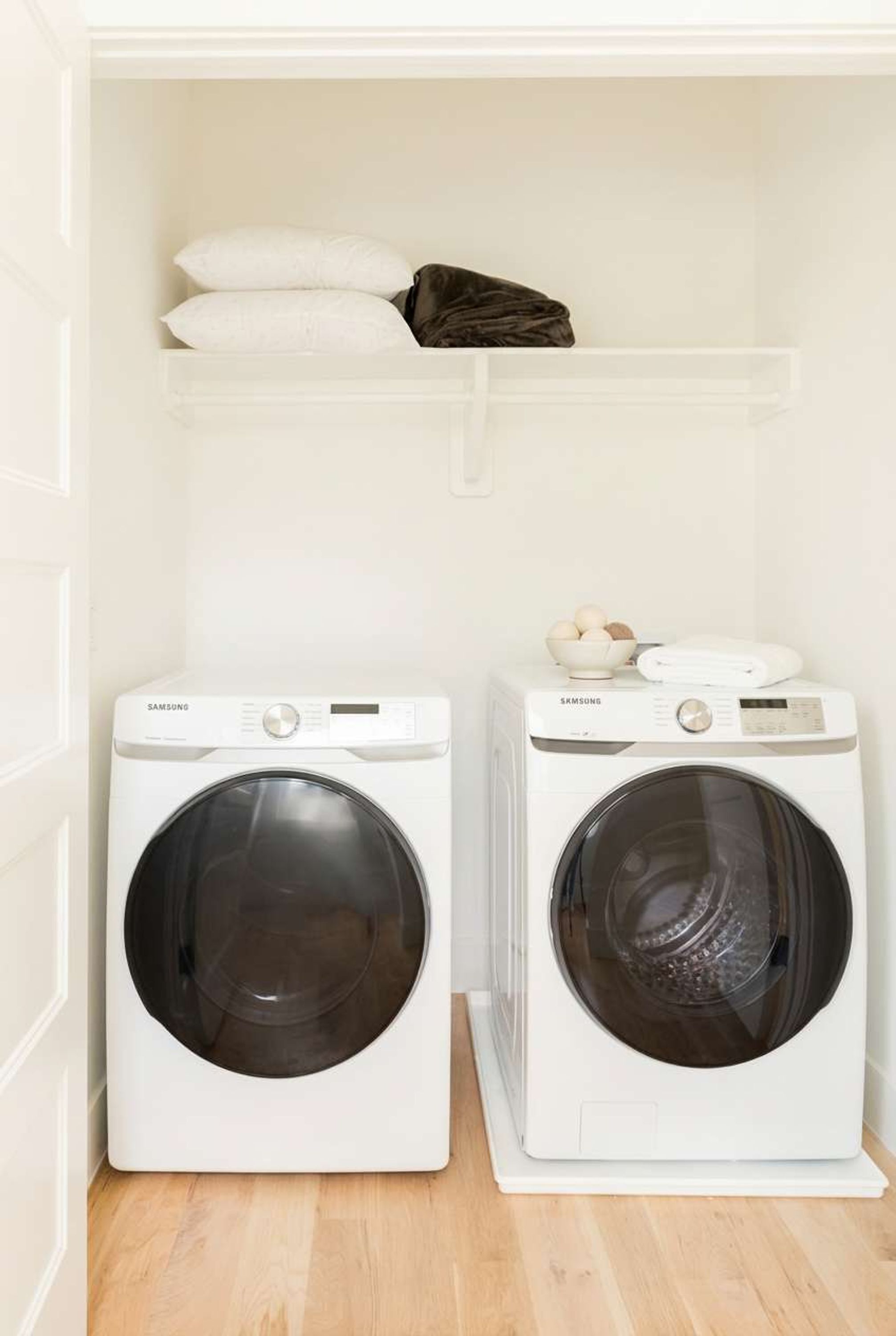 Deux lave-linge Samsung à chargement frontal, de couleur blanche, se trouvent dans une buanderie. Le sol est en bois et une étagère blanche est placée au-dessus des machines.