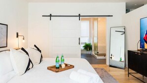 A modern hotel room with a large bed, a wooden bedside table with two bottles and a tray, a mirror, and a TV on a black cabinet. - Nash Chic Near DT Rooftop Luxe Comfort (Nashville)