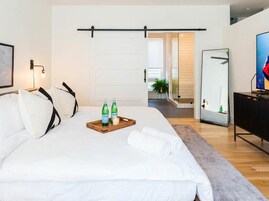 A modern hotel room with a large bed, a wooden bedside table with two bottles and a tray, a mirror, and a TV on a black cabinet.