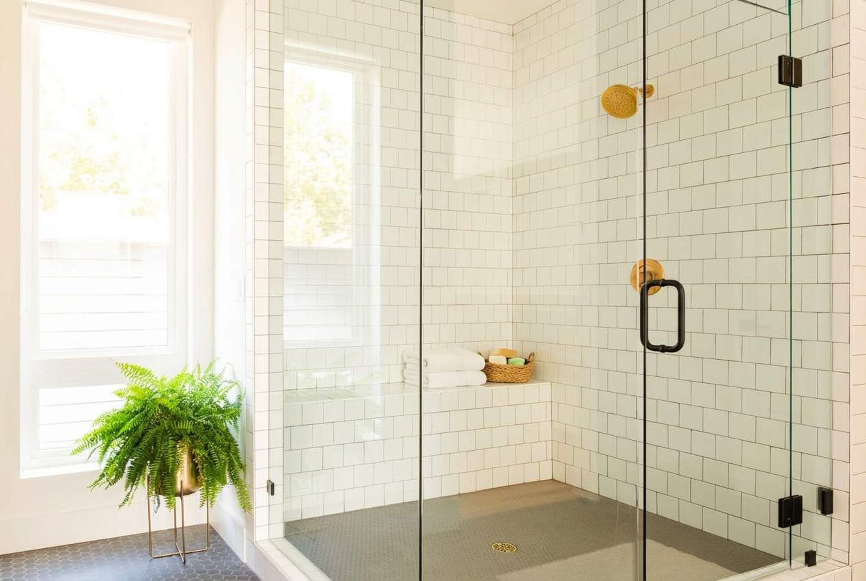 A glass-enclosed shower area with white tiles, a plant, and a basket of toiletries.