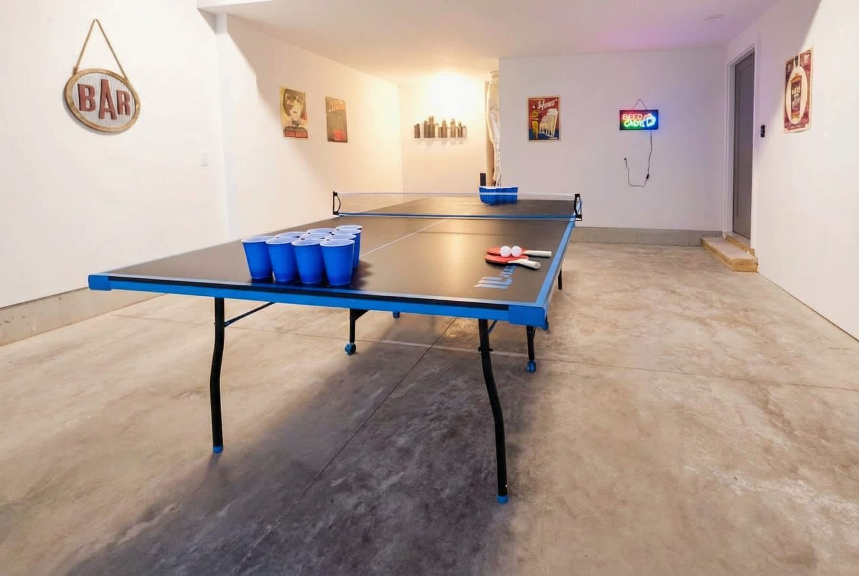 Une table de ping-pong avec des pieds bleus, un filet, ainsi que plusieurs raquettes et gobelets bleus.