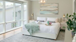 A bedroom with a large window, a bed with white bedding, a green throw blanket, and a painting on the wall.