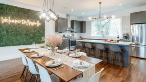 A modern kitchen with a central island, a dining area with a wooden table, and a neon sign on the wall.