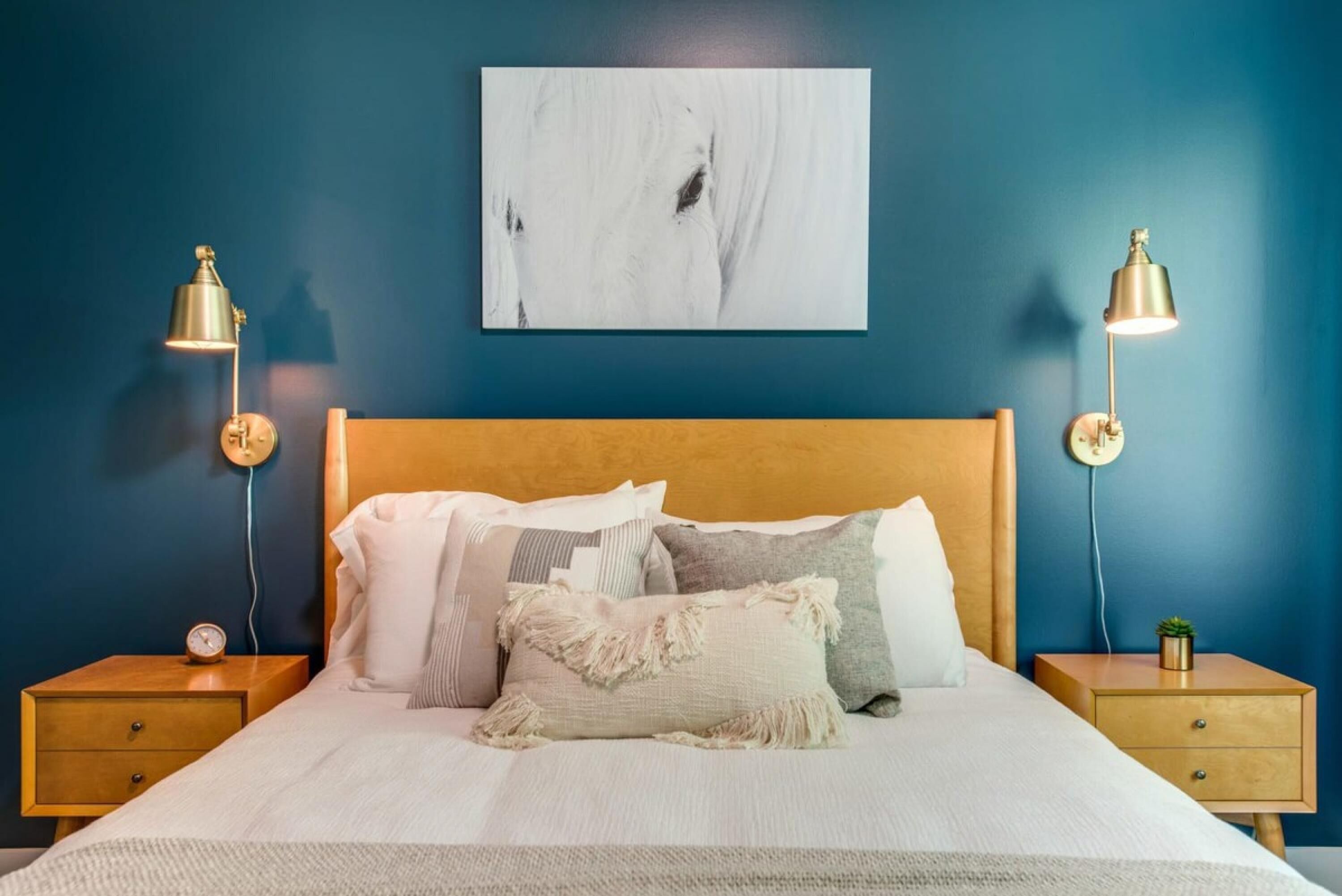 A bedroom with a wooden headboard, a white bedspread, multiple pillows, two bedside tables with lamps, and a wall-mounted abstract painting.