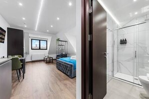 Quarto de hotel moderno com piso de madeira, uma cama, uma escrivaninha com cadeira, uma janela e box de vidro para o chuveiro.