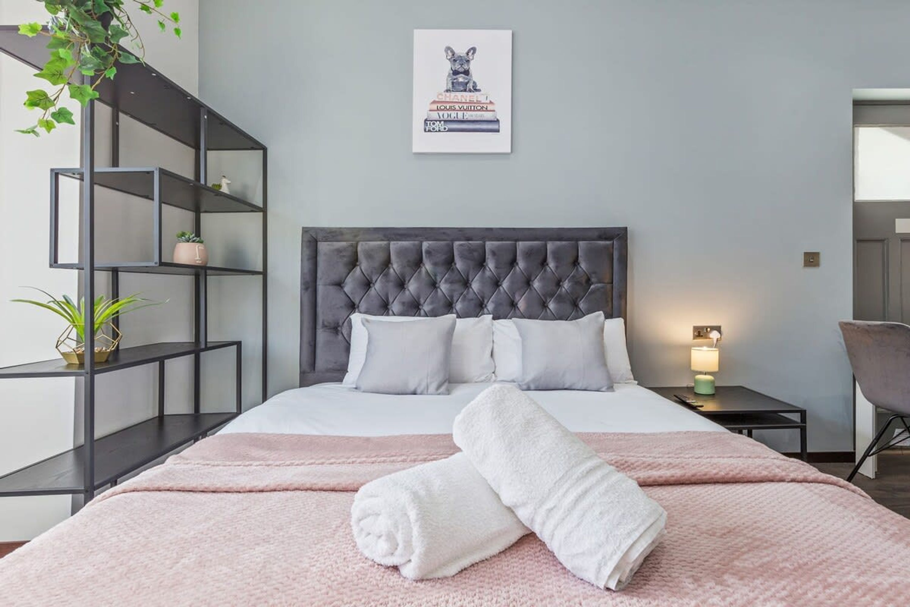 A neatly made bed with a tufted headboard, a bedside table with a lamp, and a wall-mounted picture of a dog on a stack of designer brand books.