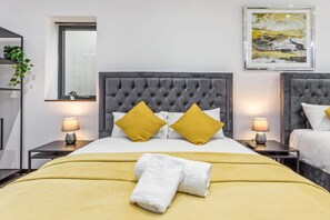 A hotel room with a tufted headboard, two bedside tables with lamps, a bed with a yellow blanket and white towels, a mirror, and a framed abstract painting on the wall. - Luxury Studio Suite Near Euston Central London (London)