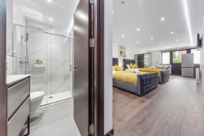 A modern hotel room with a glass shower enclosure, a large bed with yellow pillows, and a flat-screen TV mounted on the wall. - Luxury Studio Suite Near Euston Central London (London)
