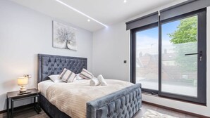 Ein modernes Schlafzimmer mit einem großen Bett, einem Nachttisch, einer Lampe, einem Fenster mit Jalousien und einem Gemälde an der Wand.