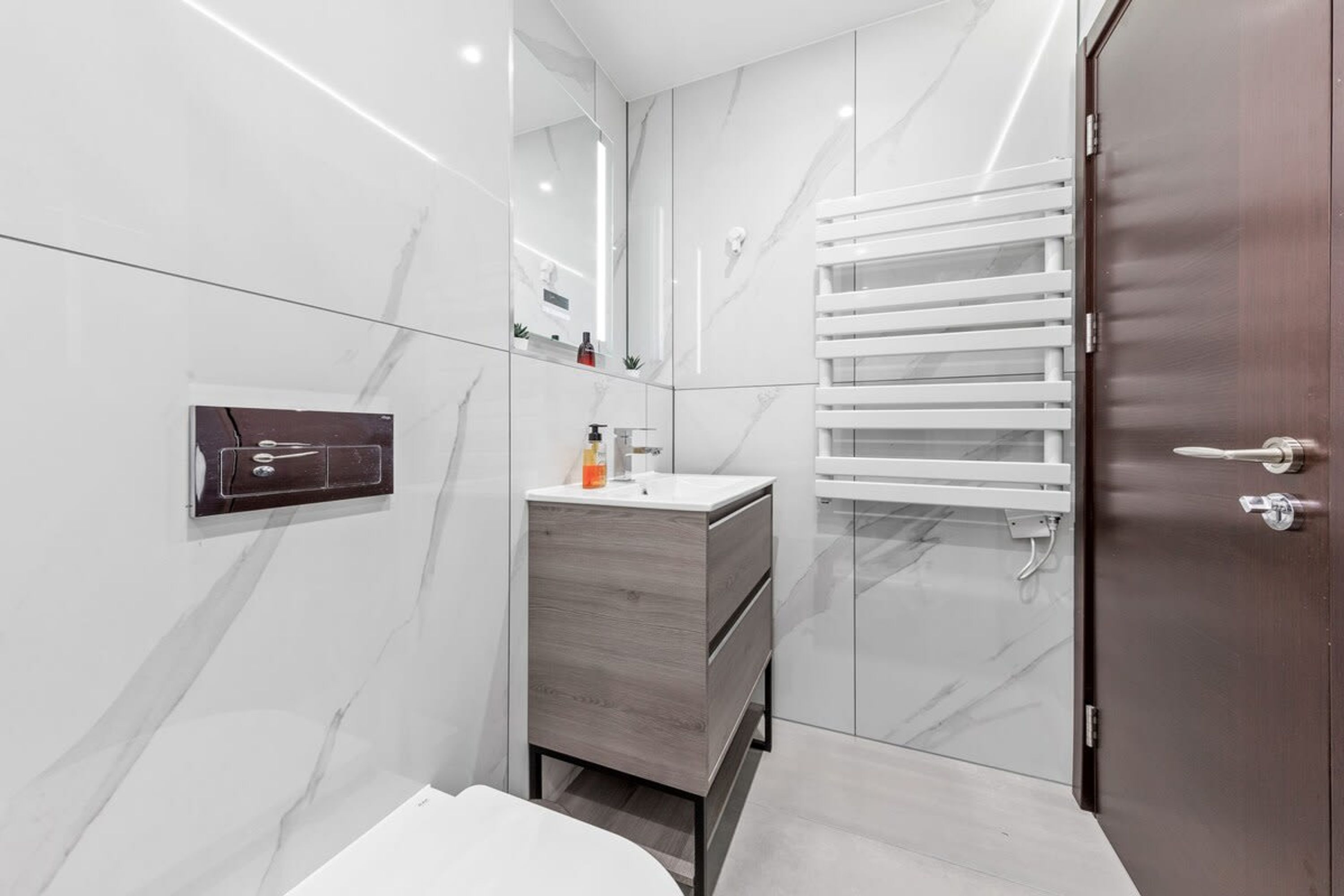 A modern bathroom with white marble walls, a wooden vanity, a towel rack, and a door.