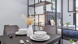 Un'area pranzo moderna con un tavolo in legno scuro apparecchiato per due persone, con ciotole e posate bianche. Sul tavolo c'è un vaso bianco con fiori secchi, e sullo sfondo un mobiletto a mensole in metallo nero con oggetti decorativi.