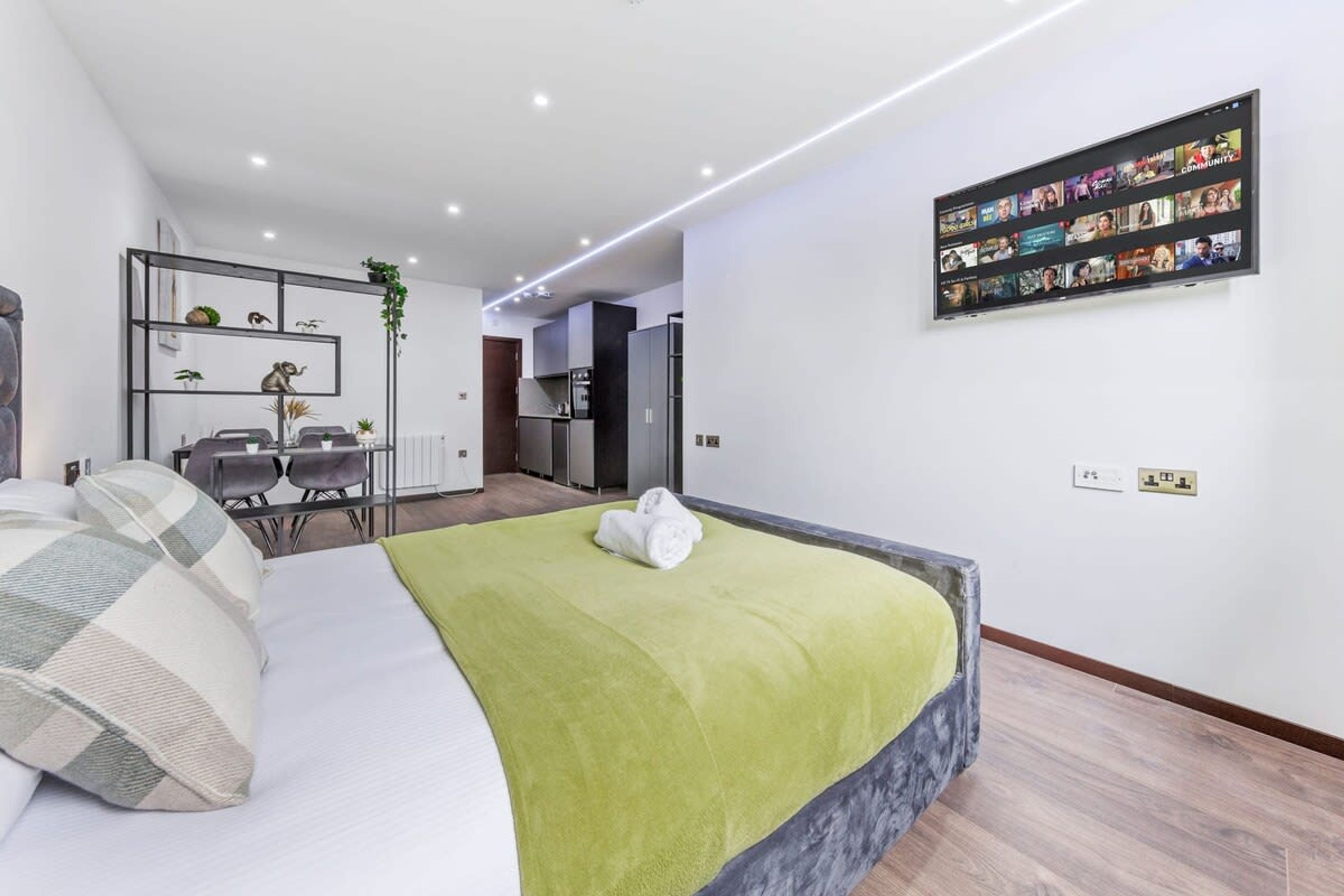 A modern hotel room with a bed, a dining area, and a kitchen in the background.