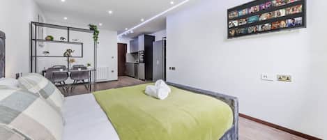 A modern hotel room with a bed, a dining area, and a kitchen in the background.