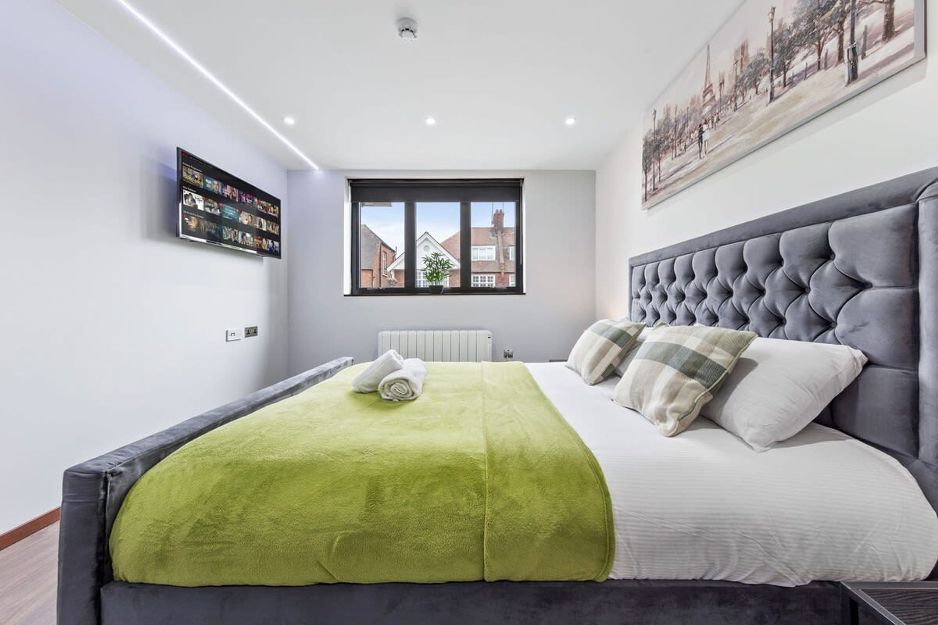 A modern bedroom with a large bed, a window with a view of buildings, a flat-screen TV on the wall, and a painting above the bed.