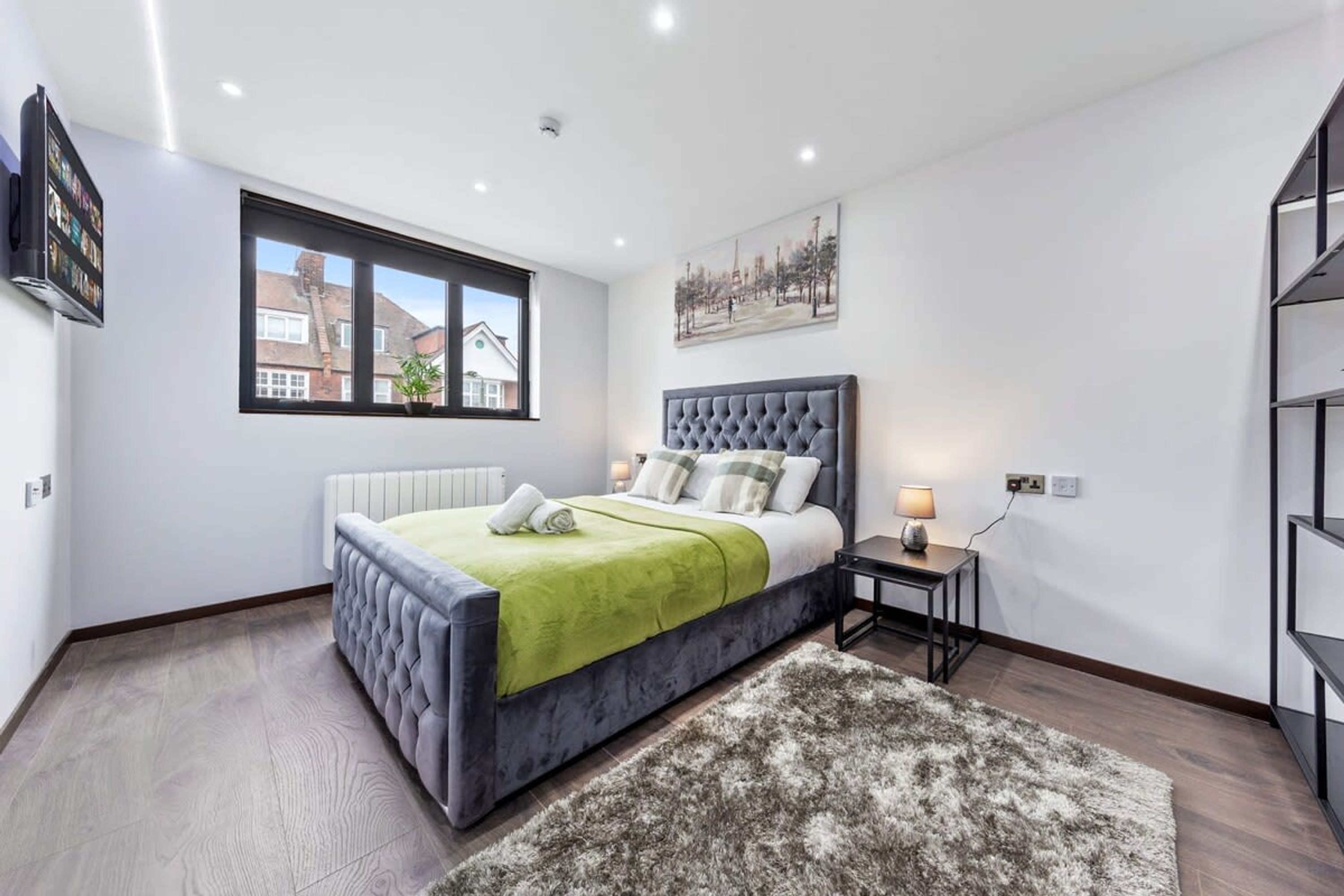 A modern bedroom with a tufted headboard, a large window, a radiator, and a bedside table with a lamp.