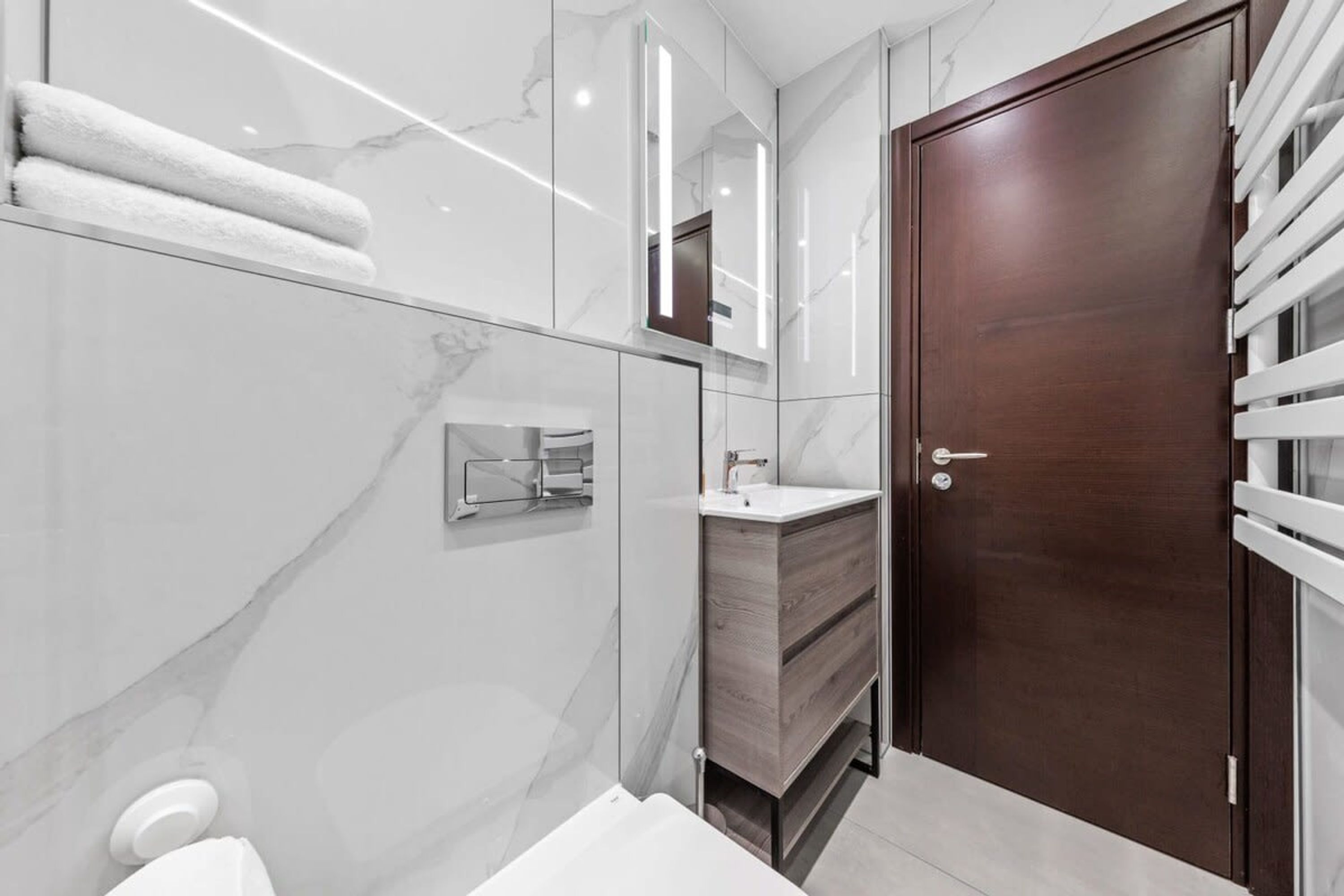 A modern bathroom with a marble wall, a wooden vanity, a white sink, and a wooden door.