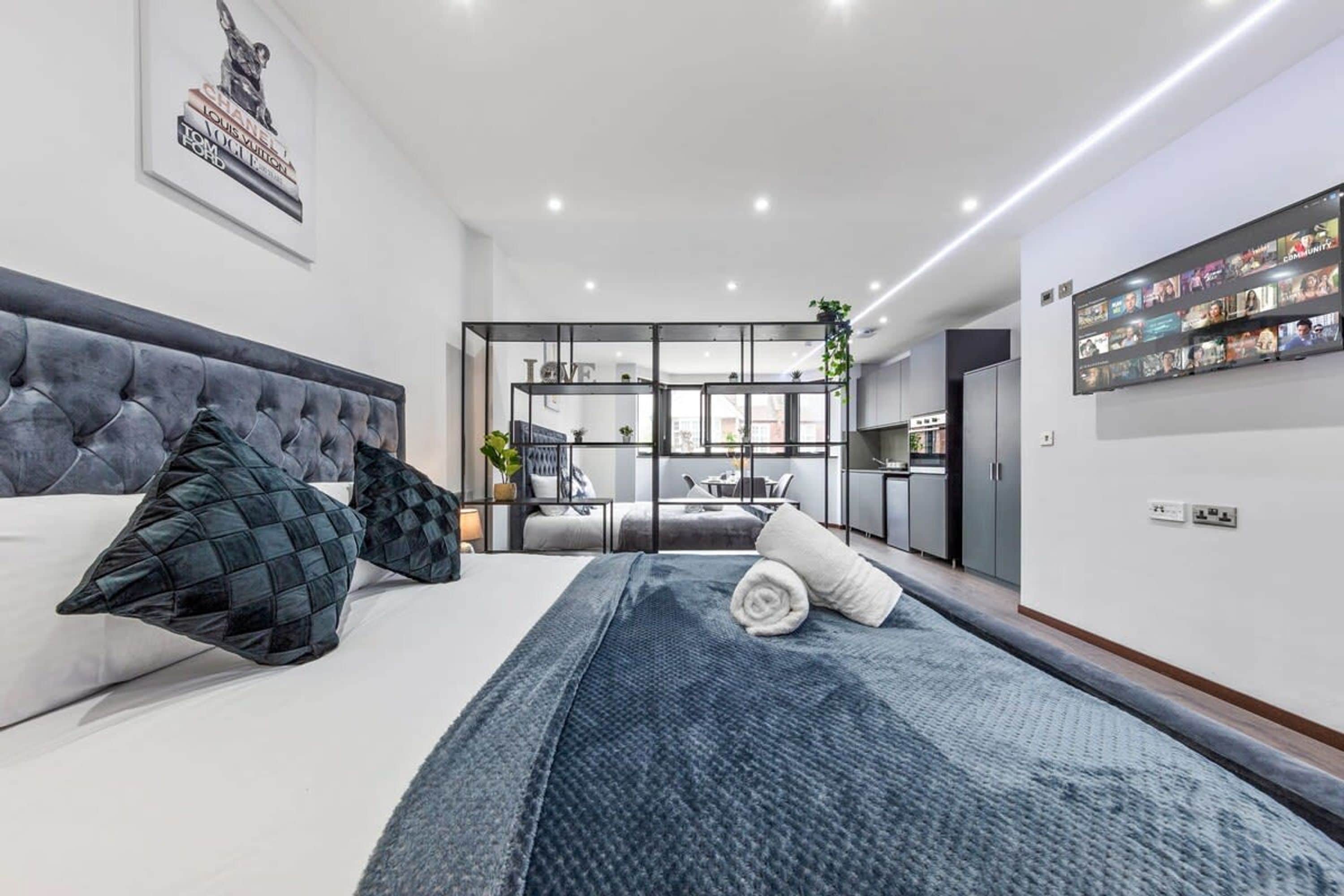 A modern hotel room with a large bed, a seating area, and a kitchen in the background.
