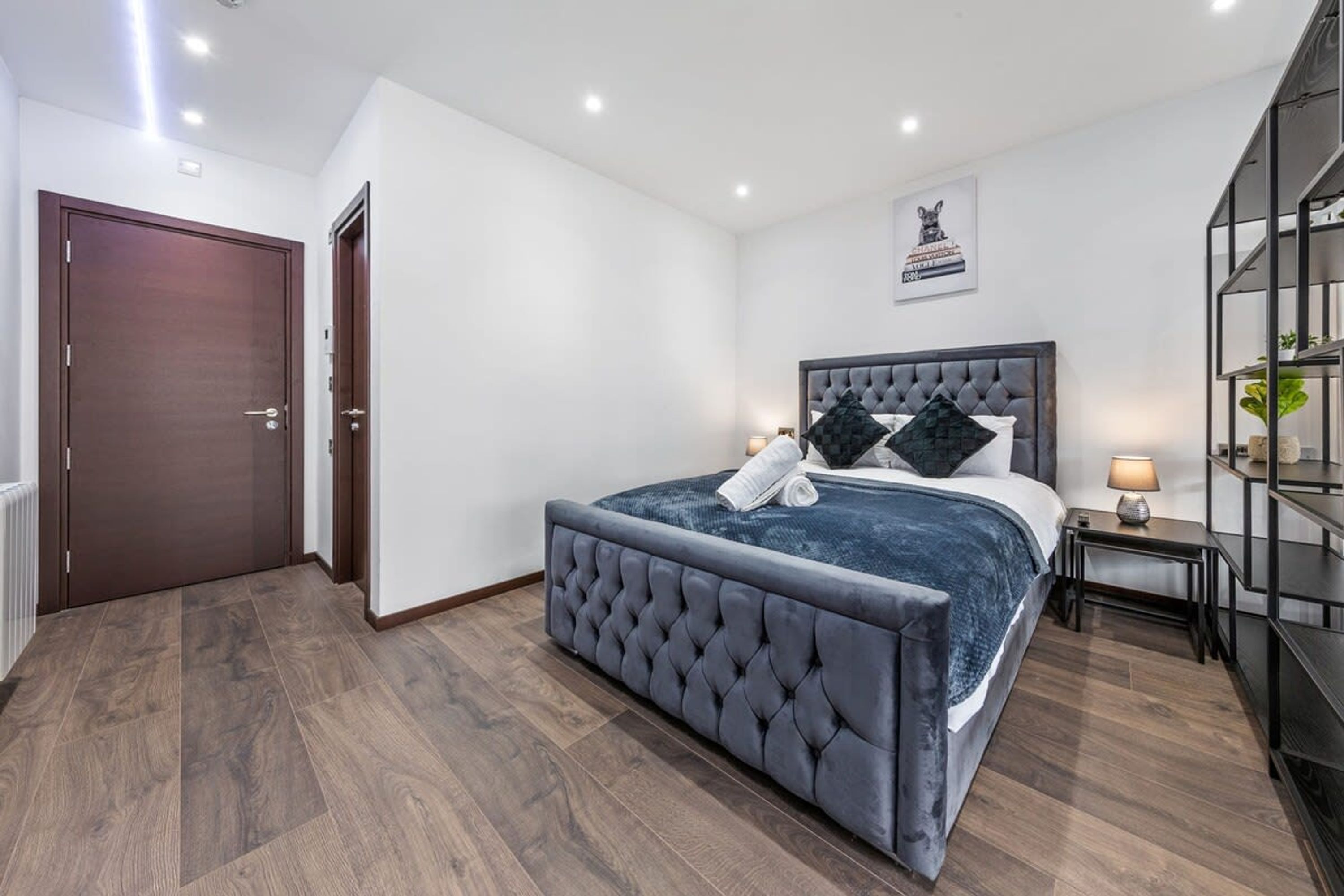 A bedroom with a large bed, a wooden floor, a dark door, and a wall-mounted shelf.