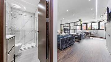 A modern bathroom with a glass shower enclosure and a separate toilet area. The living area features a large sofa, a coffee table, and a TV mounted on the wall.