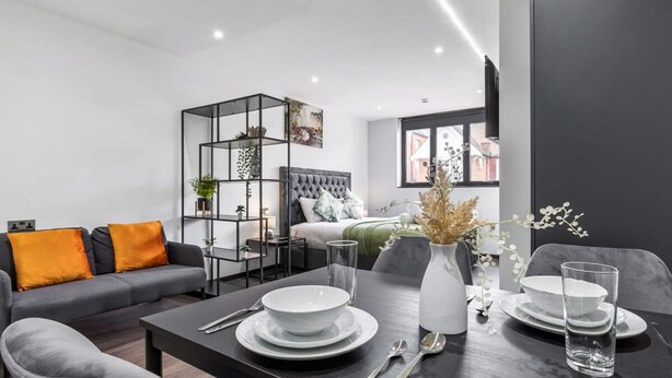 Ein modernes Wohnzimmer mit einem Esstisch für zwei Personen, einem grauen Sofa mit orangefarbenen Kissen, einer schwarzen Regalwand mit Pflanzen und einem Bett mit grauem Kopfteil.