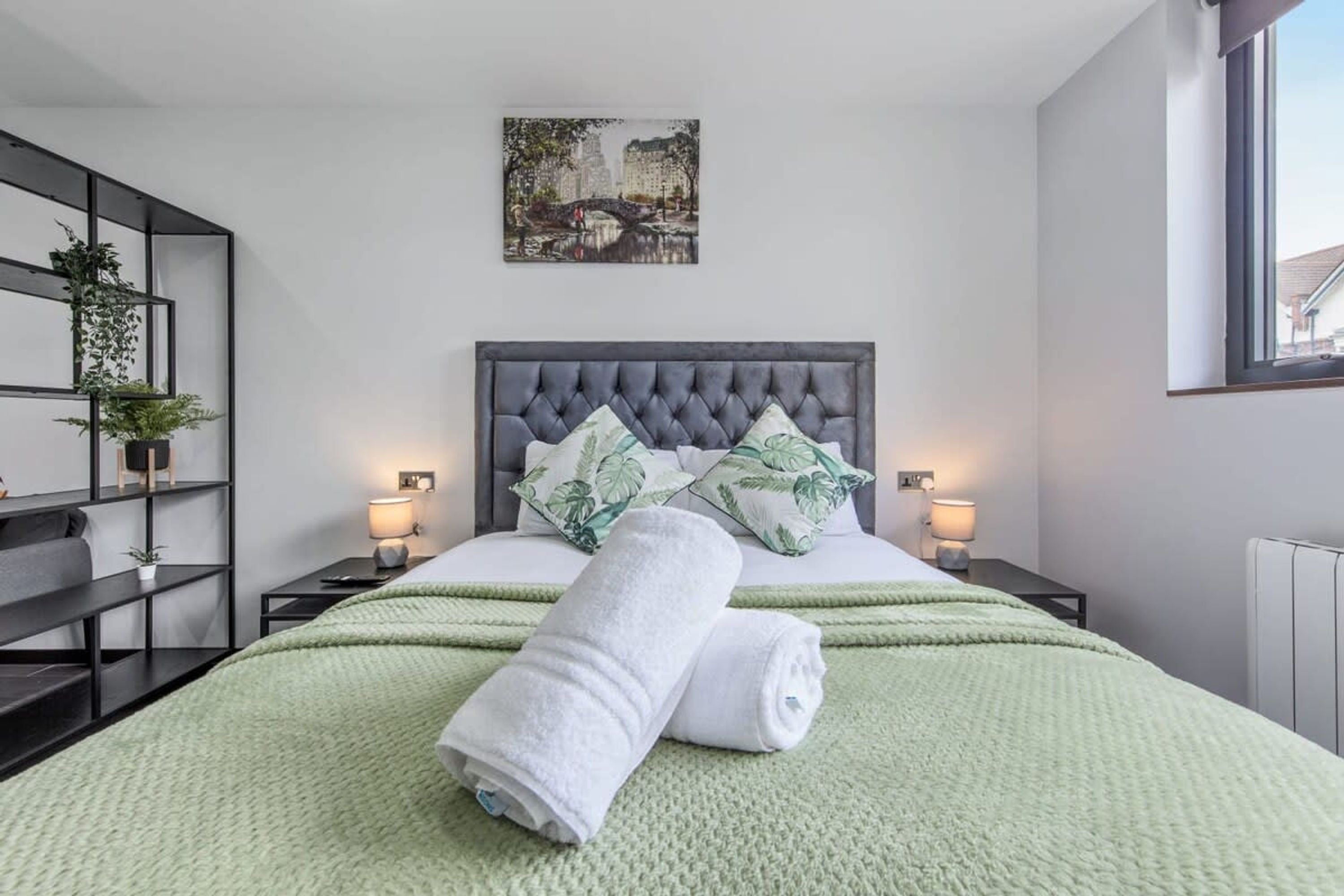 A modern bedroom with a tufted headboard, green bedding, and a cityscape painting above the bed.
