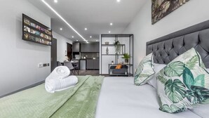 Ein modernes Hotelzimmer mit einem großen Bett, einem flachbildfernseher an der Wand, einer kleinen Küchenzeile und einer Sitzecke mit Sofa und Beistelltisch.