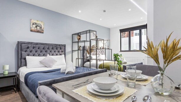 Quarto moderno com cama de cabeceira estofada, mesa de centro de vidro e área de jantar com um vaso de plantas secas.