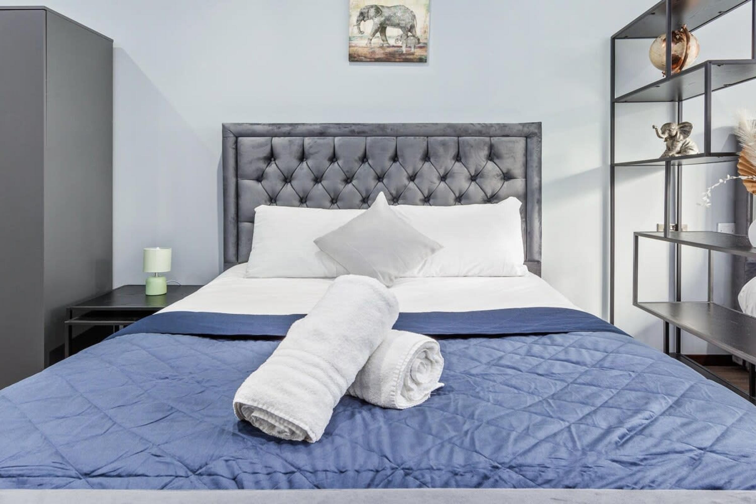 A neatly made bed with a blue quilt and white pillows, a grey headboard, and a bedside table with a lamp.