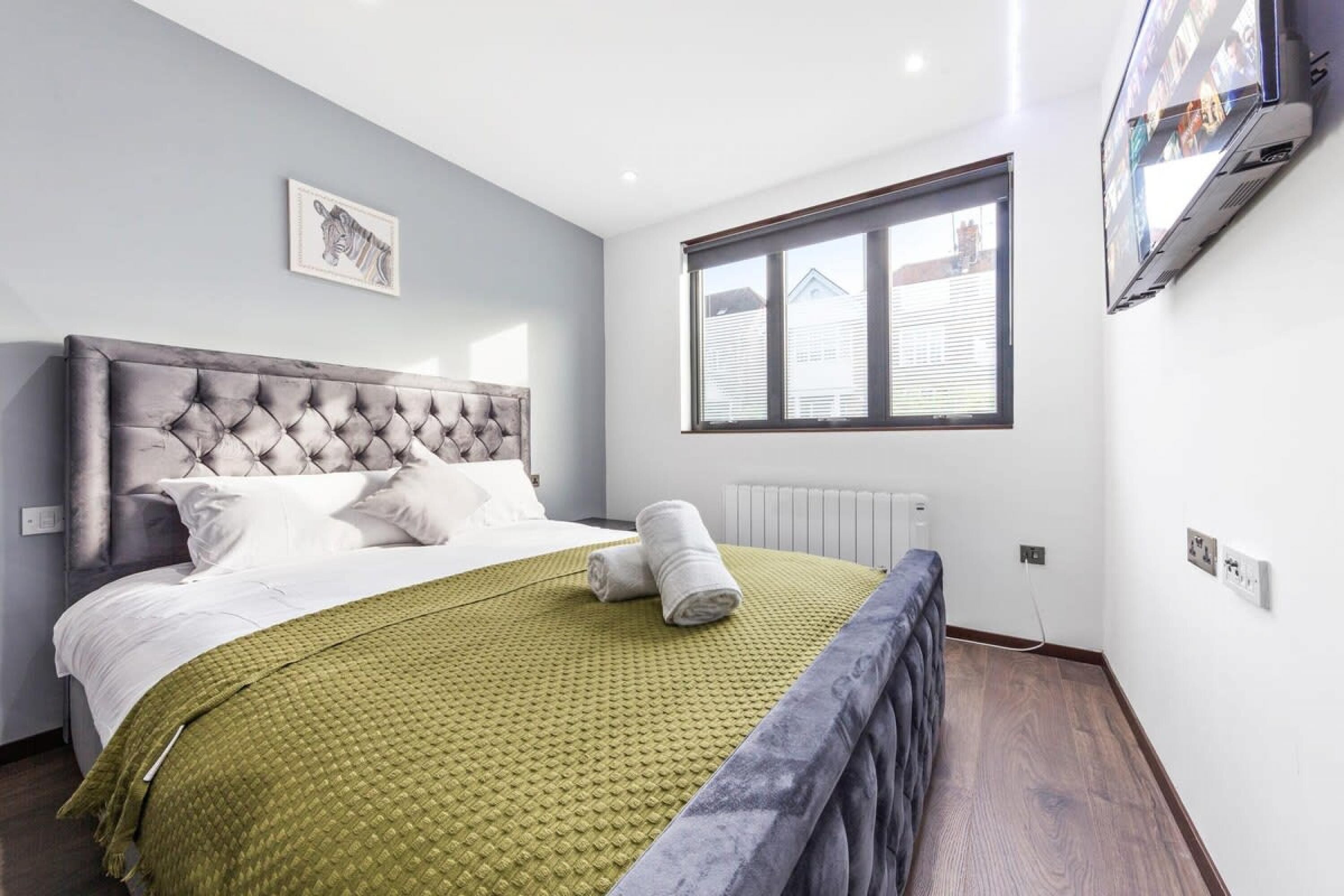 Un dormitorio con cabecera capitonada, colcha verde acolchada, toalla enrollada, ventana con persianas y un cuadro en la pared.