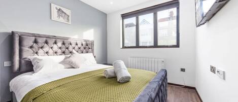 Un dormitorio con cabecera capitonada, colcha verde acolchada, toalla enrollada, ventana con persianas y un cuadro en la pared.