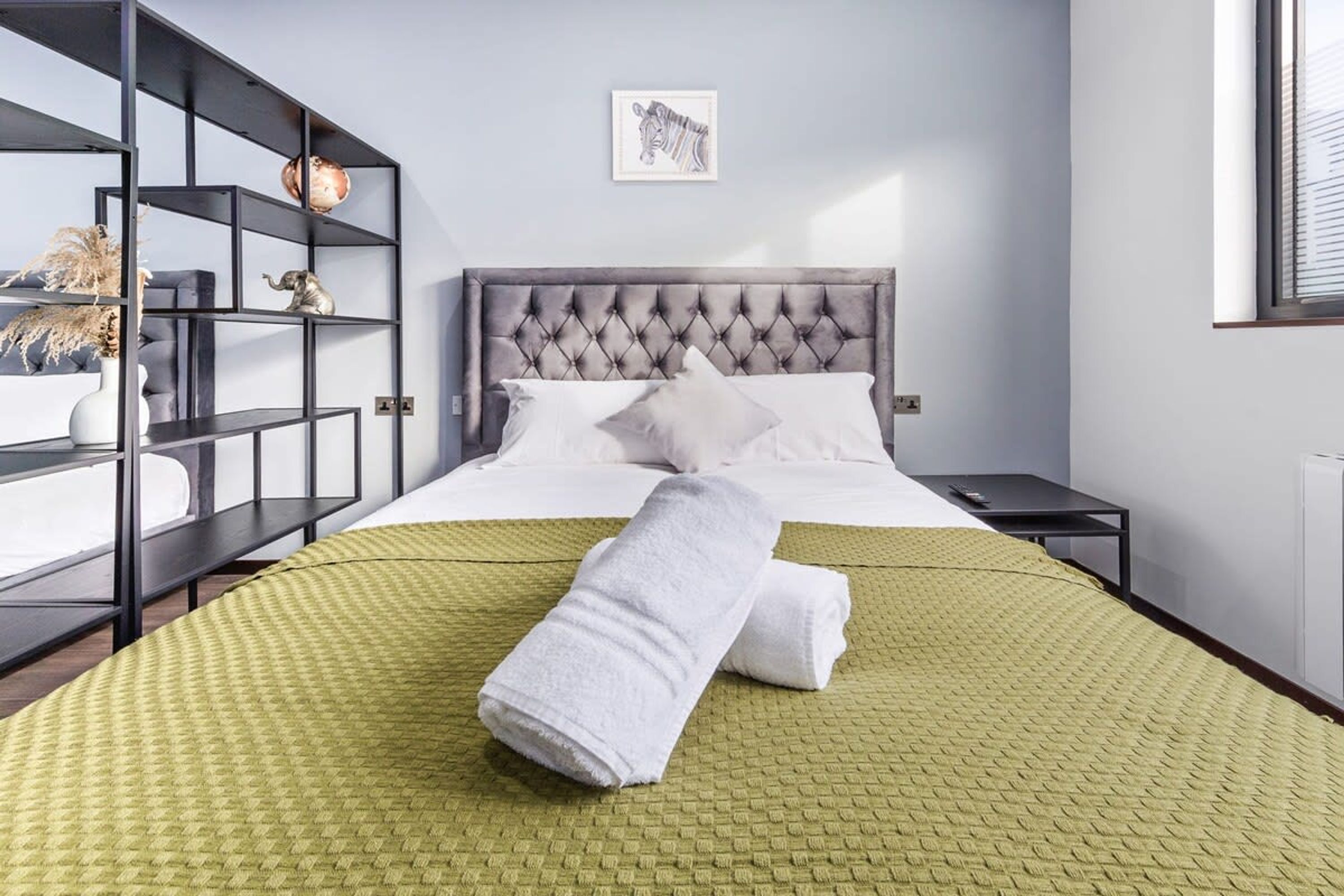 Une chambre d’hôtel avec un lit, une couvre-lit verte à motifs, une tête de lit grise à capitons, une étagère en métal noir agrémentée d’objets décoratifs et un tableau encadré au mur.