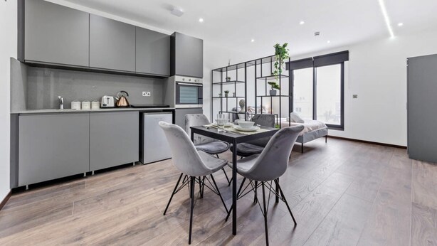 A modern kitchen with grey cabinets, a dining area with a table and chairs, and a large window providing a view of the city.