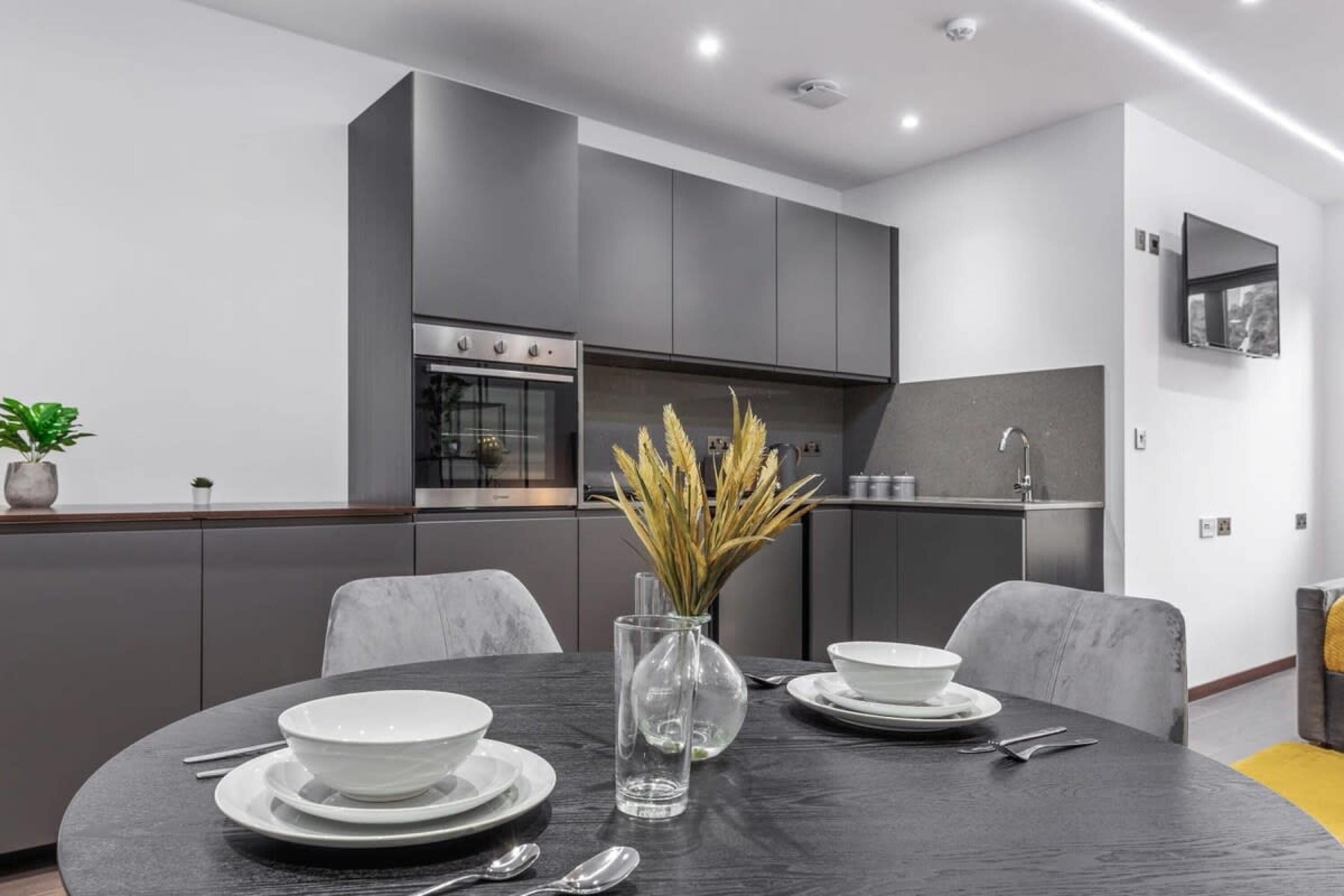 A modern kitchen with grey cabinetry, a built-in oven, and a dining area with a round table set for two.