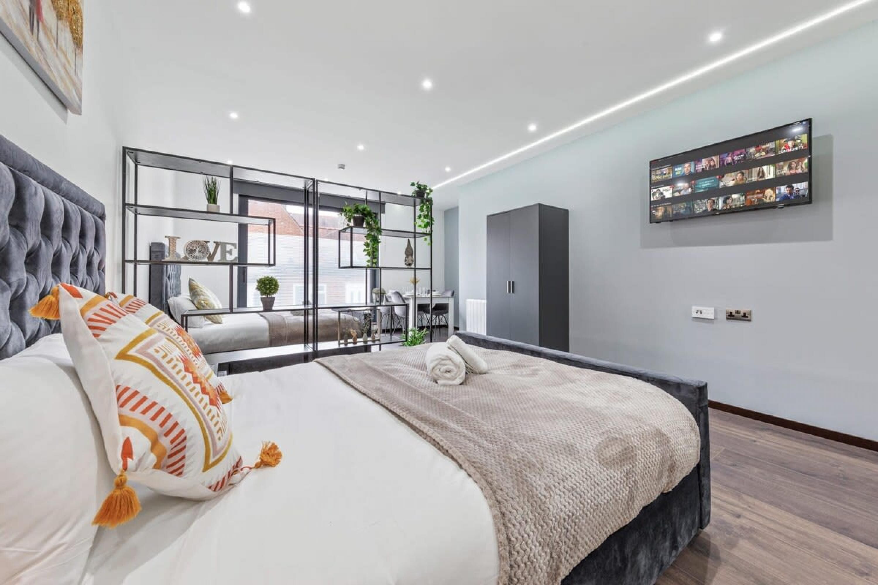 A modern bedroom with a large bed, a TV mounted on the wall, and a balcony with a view.