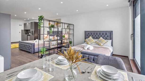 Habitación de hotel moderna con comedor, una cama con cabecera capitonada y una kitchenette al fondo.