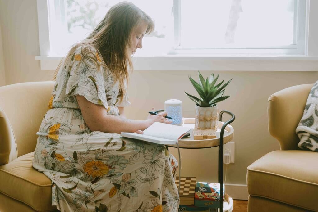 A person sitting on a beige sofa, writing in a notebook, with a small table beside them holding a plant, a candle, and a box of tissues.