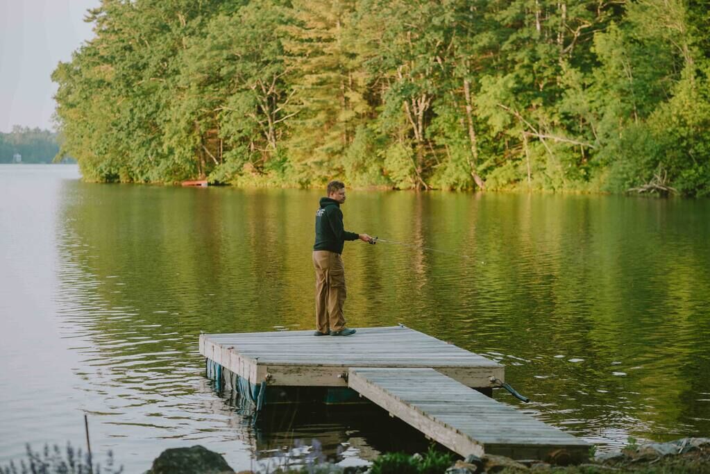 A person standing on a wooden dock extending into a calm lake, surrounded by dense green trees.