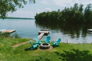 Ein Seeufer mit einer Holzterrasse, Adirondack-Stühlen, einer Feuerstelle und einem Steg, der ins Wasser führt.